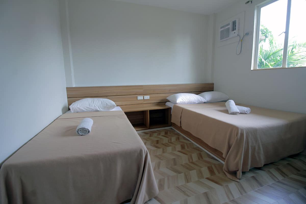 A hotel room with two single beds, a wooden headboard, a small window, and a built-in heater.