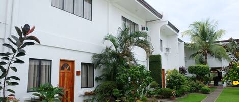 A white two-story house with a wooden door, multiple windows, and a well-maintained garden with various plants and a lawn.