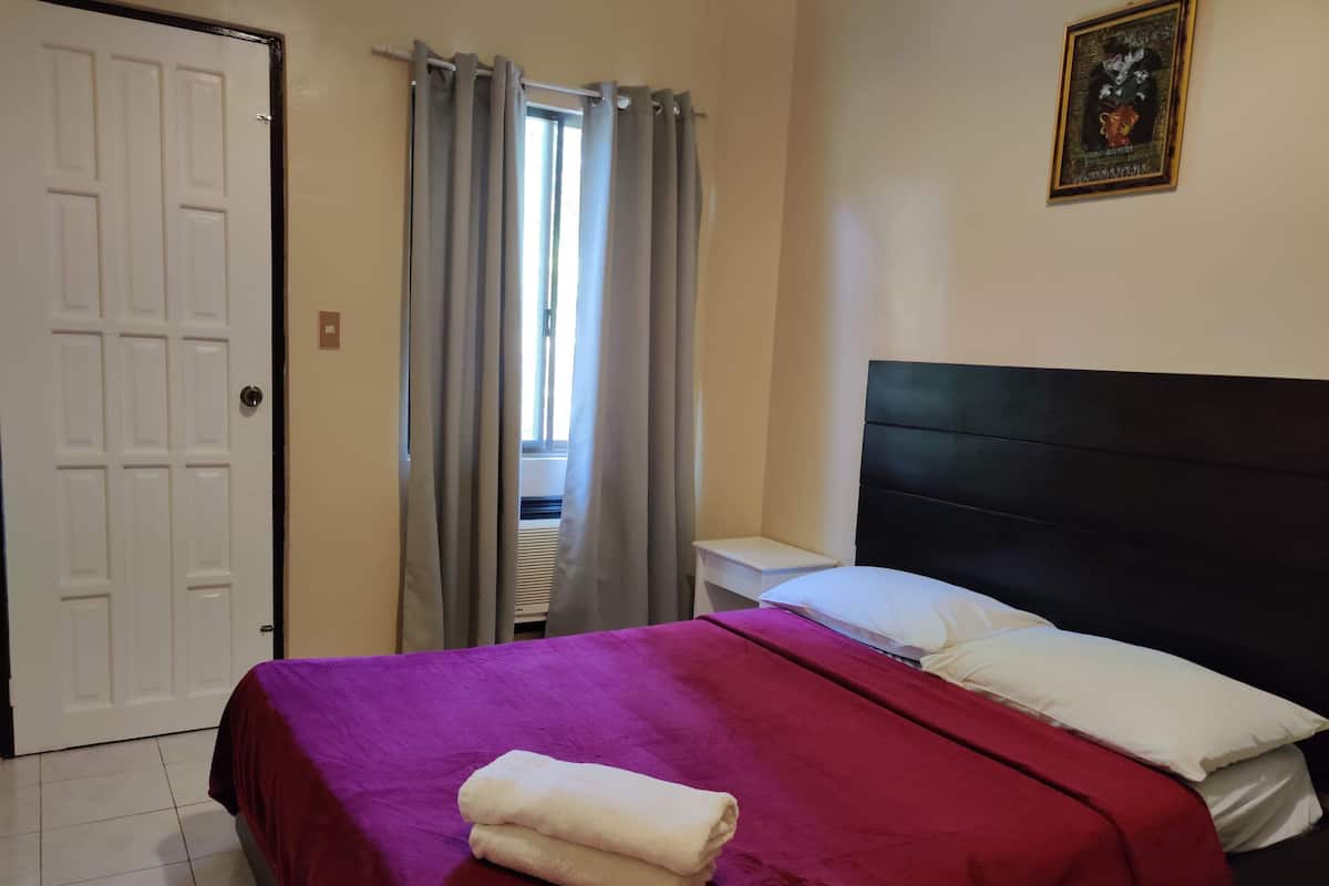 A hotel room with a bed, a nightstand, a window with curtains, and a door.