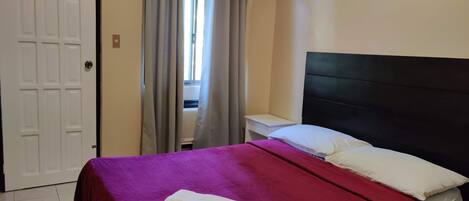 A hotel room with a bed, a nightstand, a window with curtains, and a door.