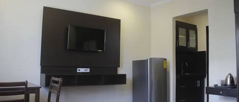 Habitación de hotel con televisión de pantalla plana montada en la pared, refrigerador, mesa de comedor con sillas y pequeña zona de kitchenette.