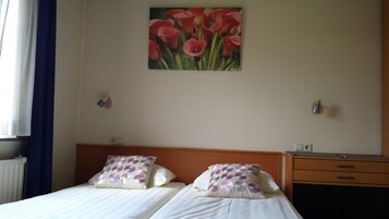 Quarto de hotel com duas camas, um quadro de flores vermelhas na parede, cabeceira de madeira e mesa de cabeceira com luminária.