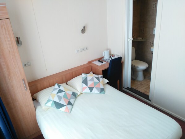 Habitación de hotel con cama, escritorio, silla, mesita de noche, cafetera y un inodoro visible a través de una puerta abierta.