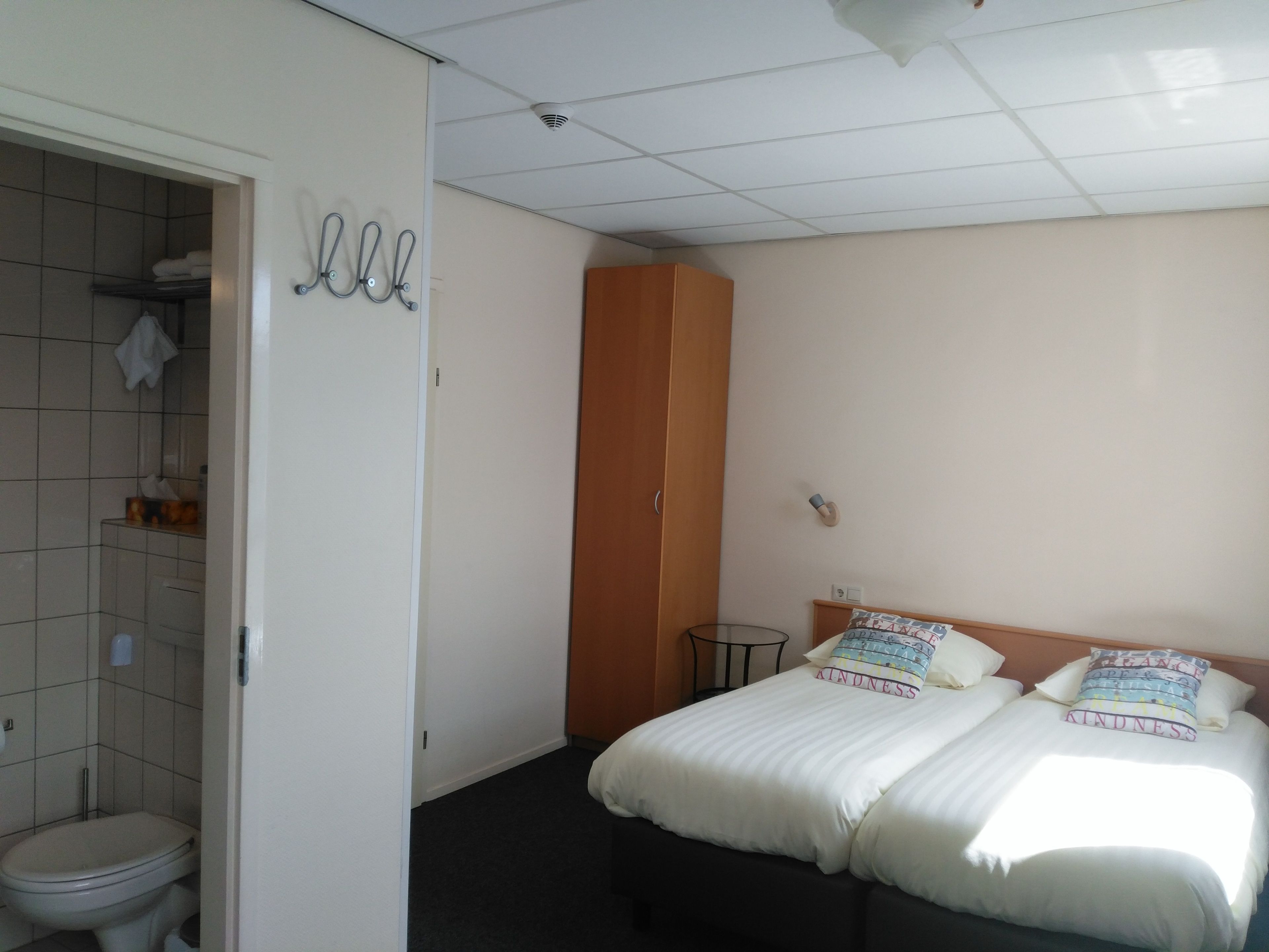 A hotel room with a bed, a wooden wardrobe, a small table, and a toilet.