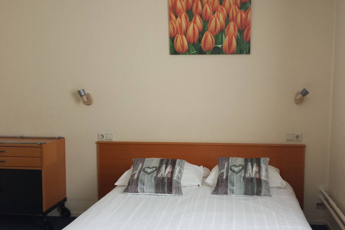 Ein Hotelzimmer mit einem Holzkopfteil, einem Bett mit weißer Bettwäsche, zwei Kissen mit Herzmuster und einem Wandbild von Tulpen sowie einem Nachttisch mit einer Lampe.