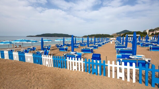 A beach with rows of blue lounge chairs and umbrellas, a white picket fence, and a hilly landscape in the background. - Bella Vista (Becici)