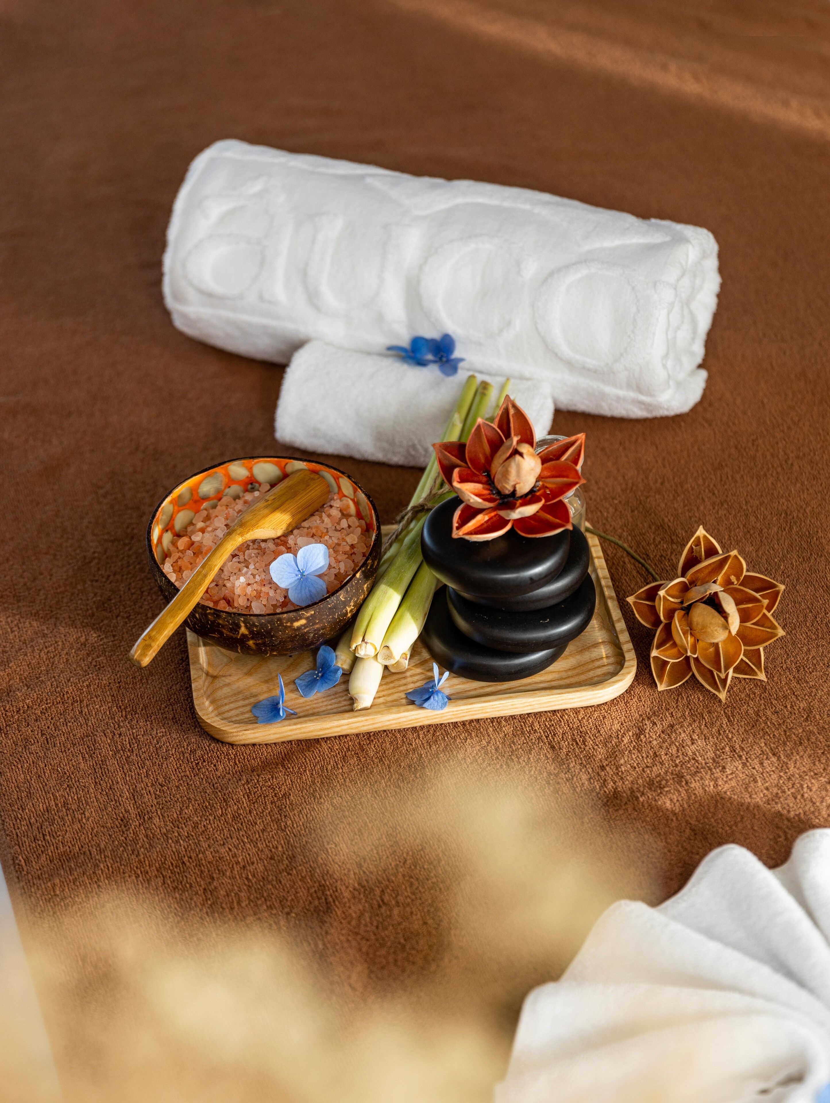 A wooden tray with a bowl of pink salt, a golden spoon, and a decorative flower.