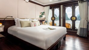 A spacious hotel room with a large bed, wooden flooring, a nightstand, a lamp, and a view of the sea through a large window with curtains. - The Au Co Cruise - Longest Cruise 2 Bays (Tuan Chau)