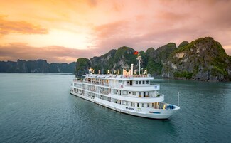 A large white river cruise ship with multiple decks and a flag, sailing on calm waters with a backdrop of green-clad limestone cliffs under a sunset sky.