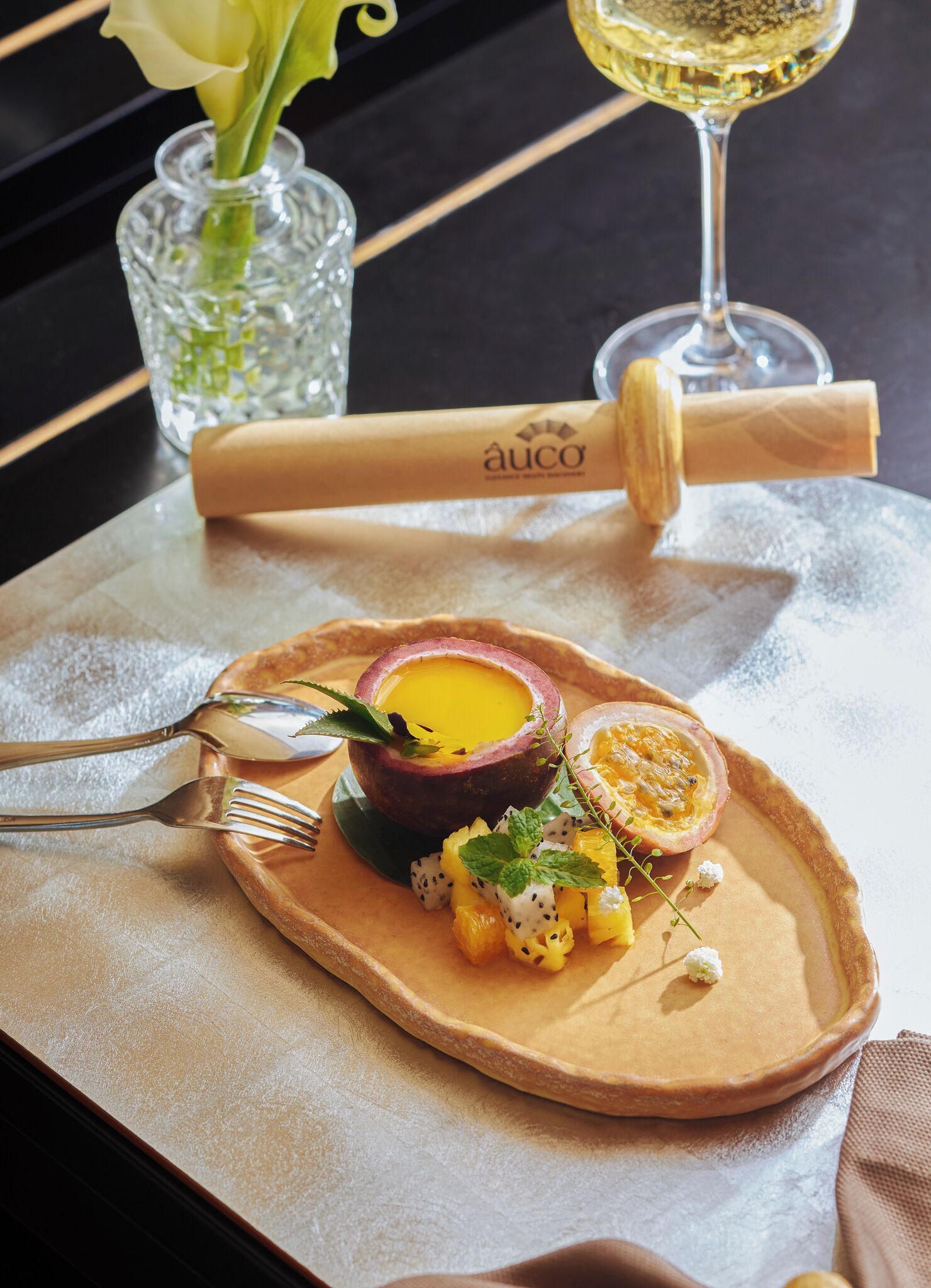 A dessert plate with a custard-like dish, passion fruit, and tropical fruits, accompanied by a wine glass and a rolled napkin on a table.