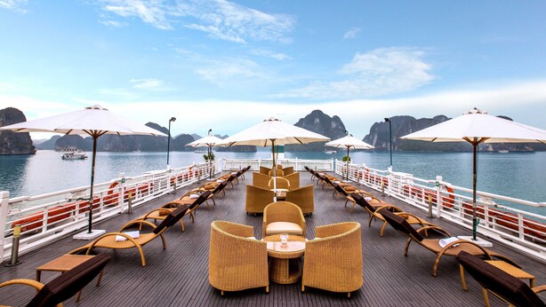 A deck with wicker furniture and umbrellas, overlooking a body of water with boats and rocky formations. - The Au Co Cruise - Longest Cruise 2 Bays (Tuan Chau)