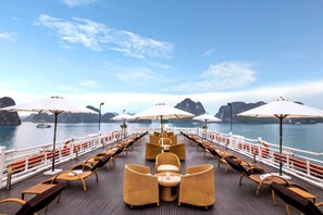A deck with wicker furniture and umbrellas, overlooking a body of water with boats and rocky formations.