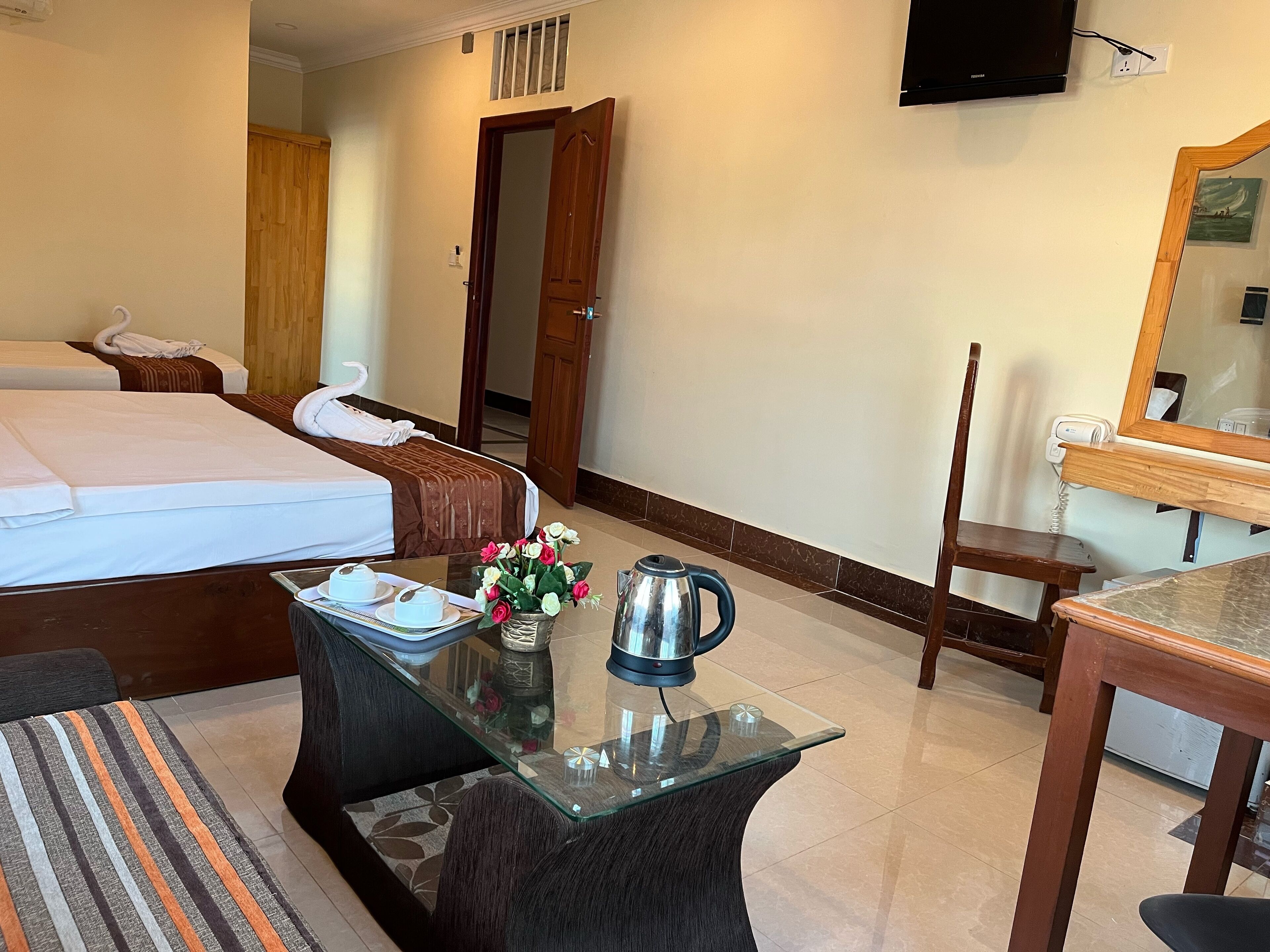 A hotel room with a bed, a glass coffee table with a kettle, a chair, and a desk with a mirror.