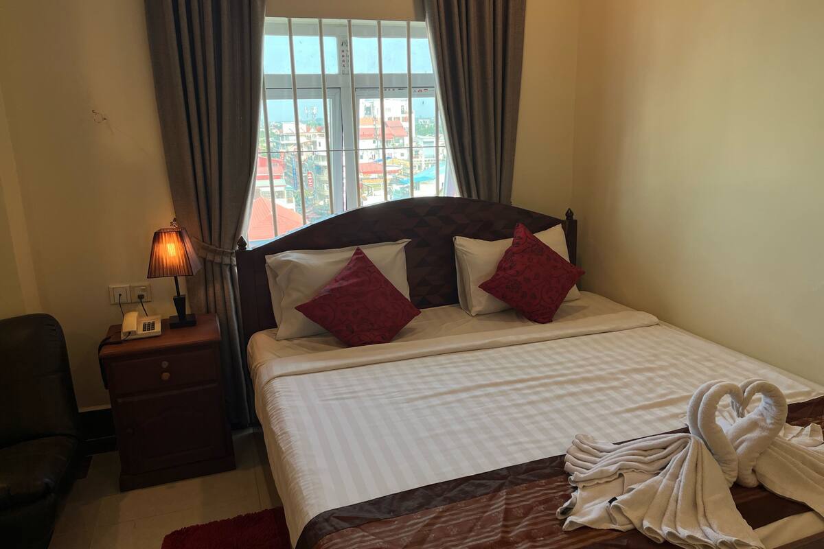 Habitación de hotel con cama, lámpara de noche, teléfono y vista a edificios fuera de la ventana.