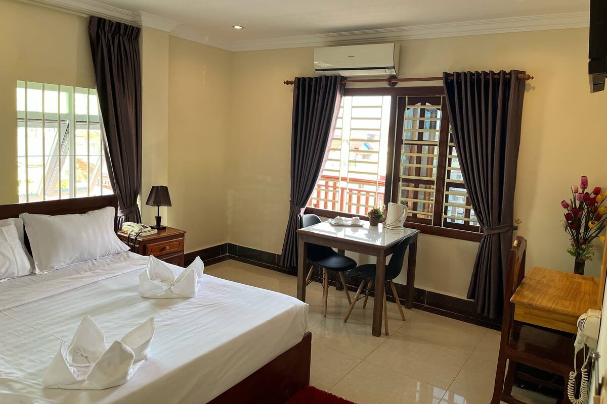 Habitación de hotel con una cama bien hecha, un escritorio con una silla, una mesita con teléfono y una ventana con cortinas.