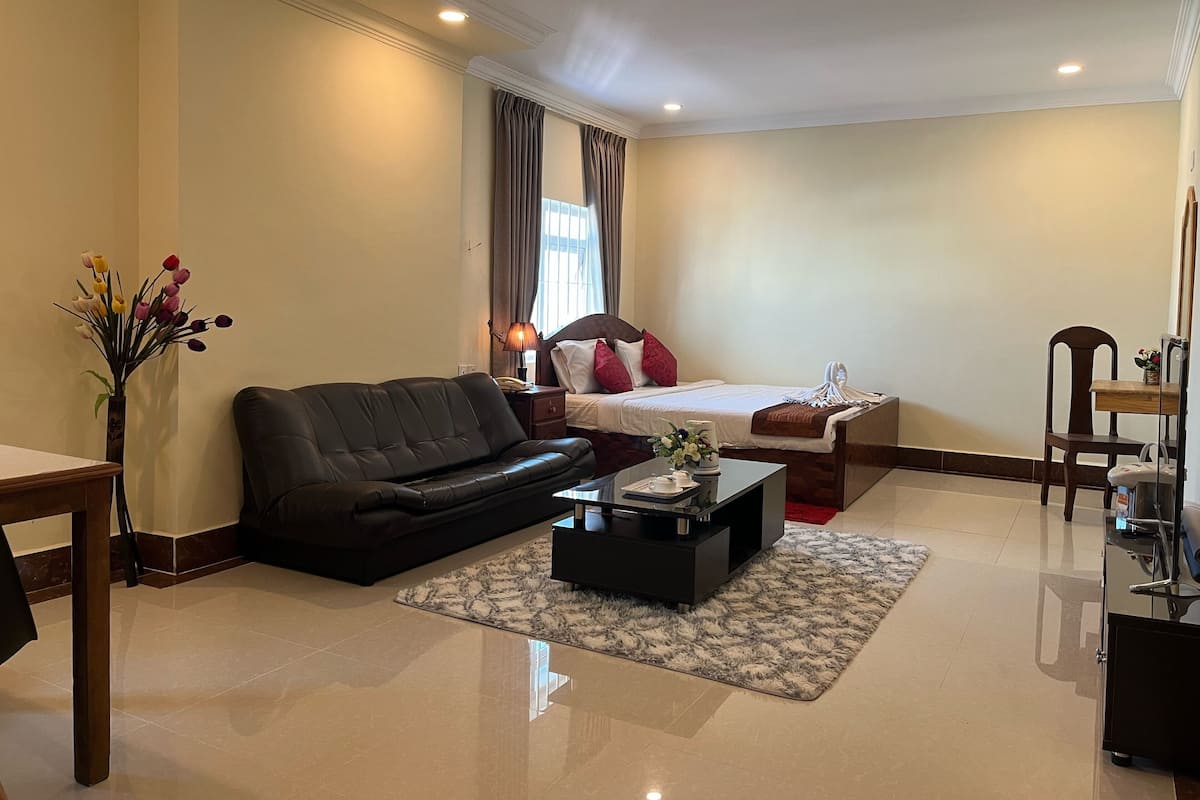 Habitación de hotel con cama, sofá de cuero negro, mesa de centro, mesita de noche con lámpara y un jarrón con flores.