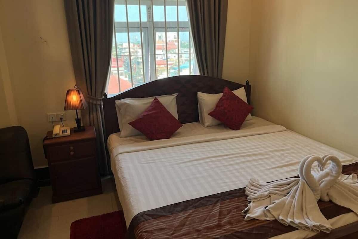 Habitación de hotel con cama, mesita de noche, lámpara y vista a edificios al exterior.