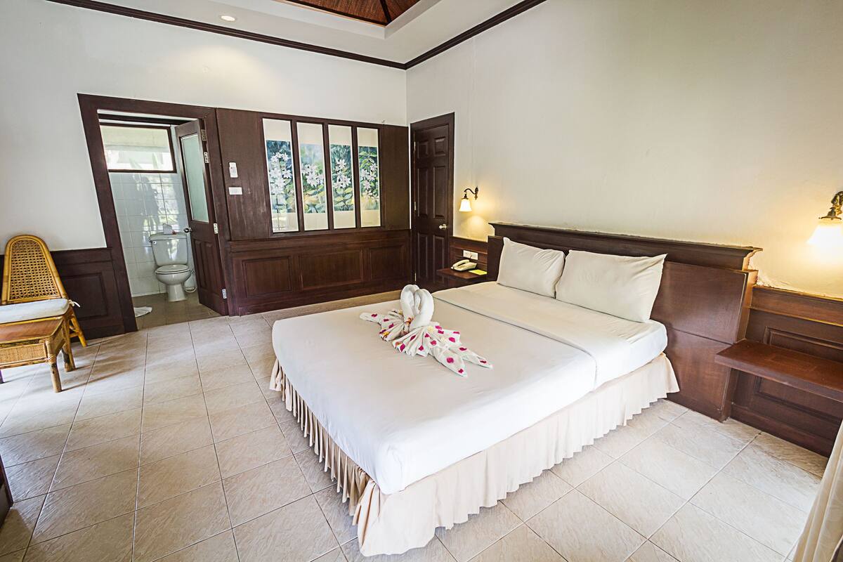 A hotel room with a large bed, a wooden headboard, a nightstand, a bedside table, a chair, and a view of the outside through a door.
