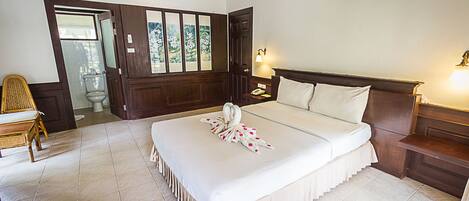 A hotel room with a large bed, a wooden headboard, a nightstand, a bedside table, a chair, and a view of the outside through a door.