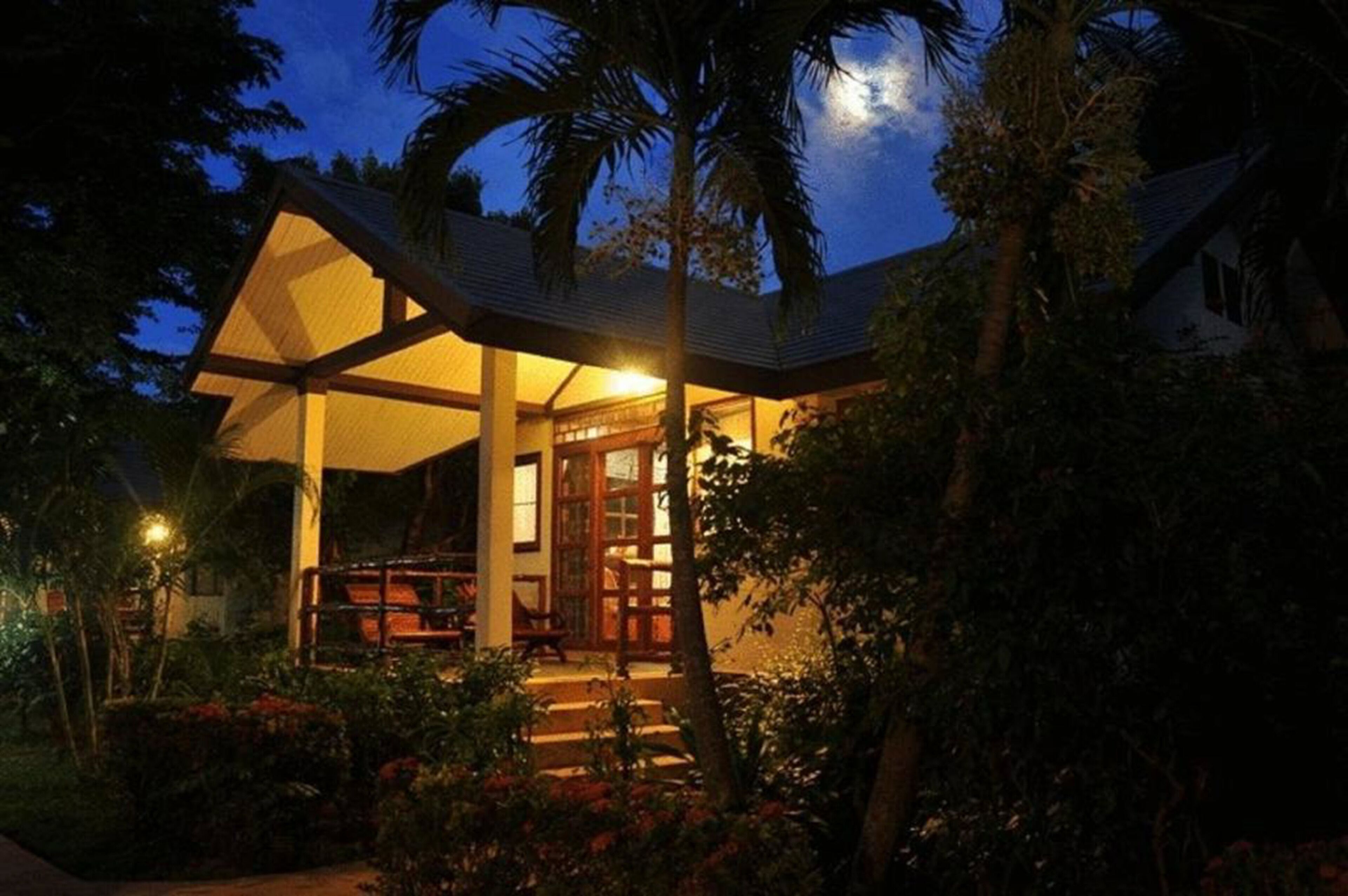 Een huis met een overdekte veranda, omringd door weelderige begroeiing en palmbomen.