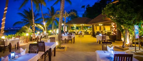 An outdoor restaurant with tables and chairs, palm trees, and a view of the ocean at dusk.