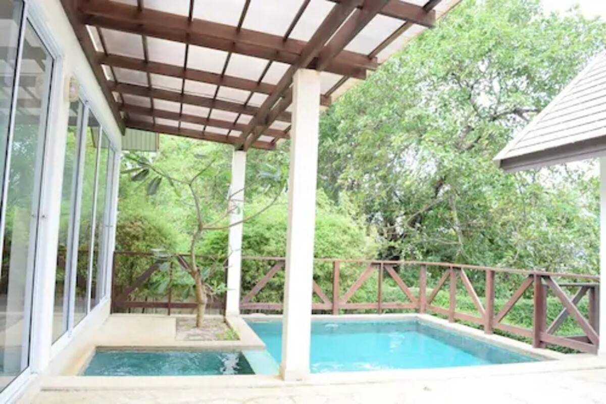 A covered outdoor swimming pool with a wooden deck and glass walls, surrounded by lush greenery.