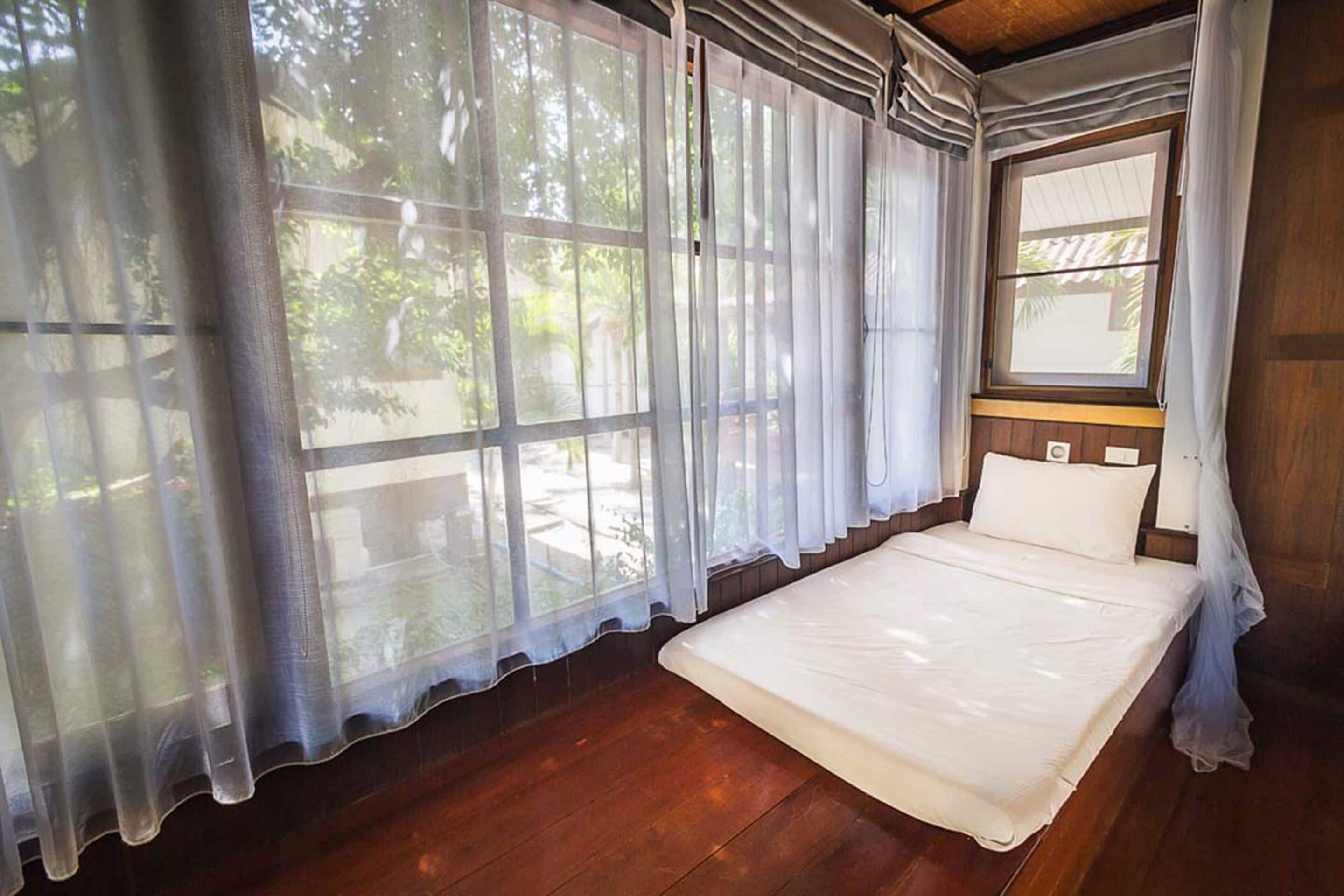 A single bed in a room with wooden flooring, large windows with sheer curtains, and a view of greenery outside.