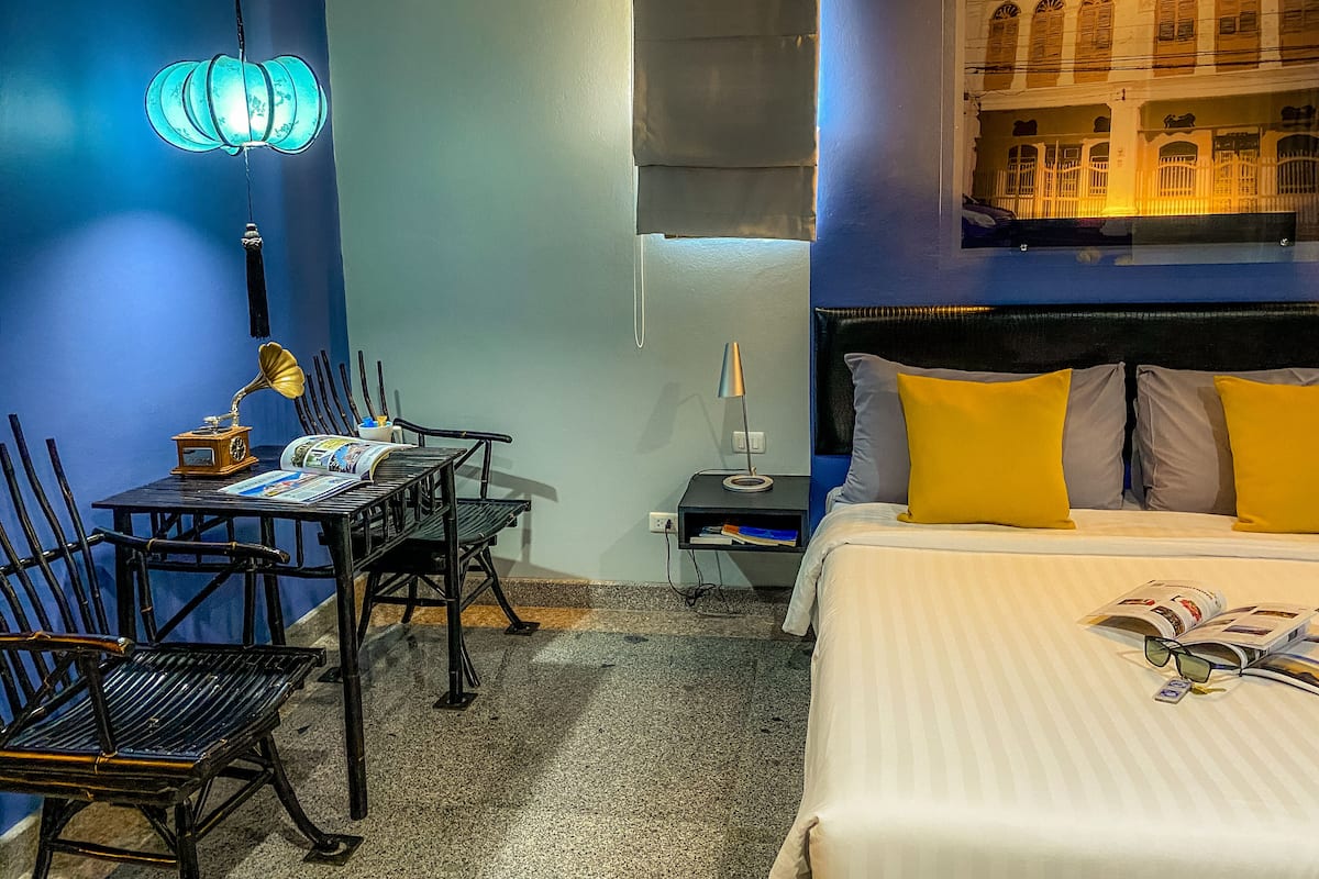 A hotel room with a bed, a desk with a lamp, a chair, and a wall-mounted light fixture.