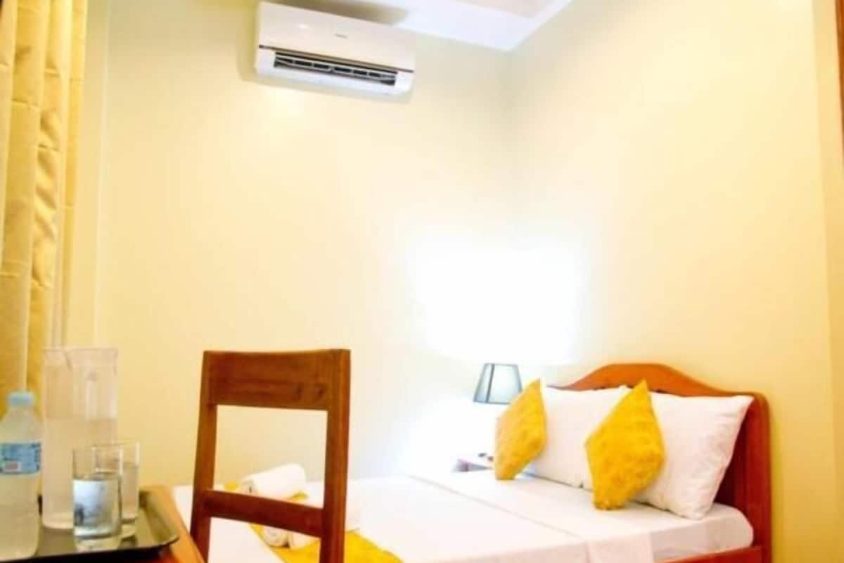 A hotel room with a bed, a chair, a bedside table with a bottle, and an air conditioning unit.