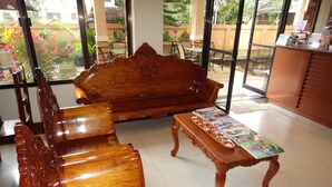 Un canapé et des fauteuils en bois se trouvent dans une pièce dotée de grandes fenêtres, d’un sol carrelé et d’une petite table sur laquelle sont posés des magazines.