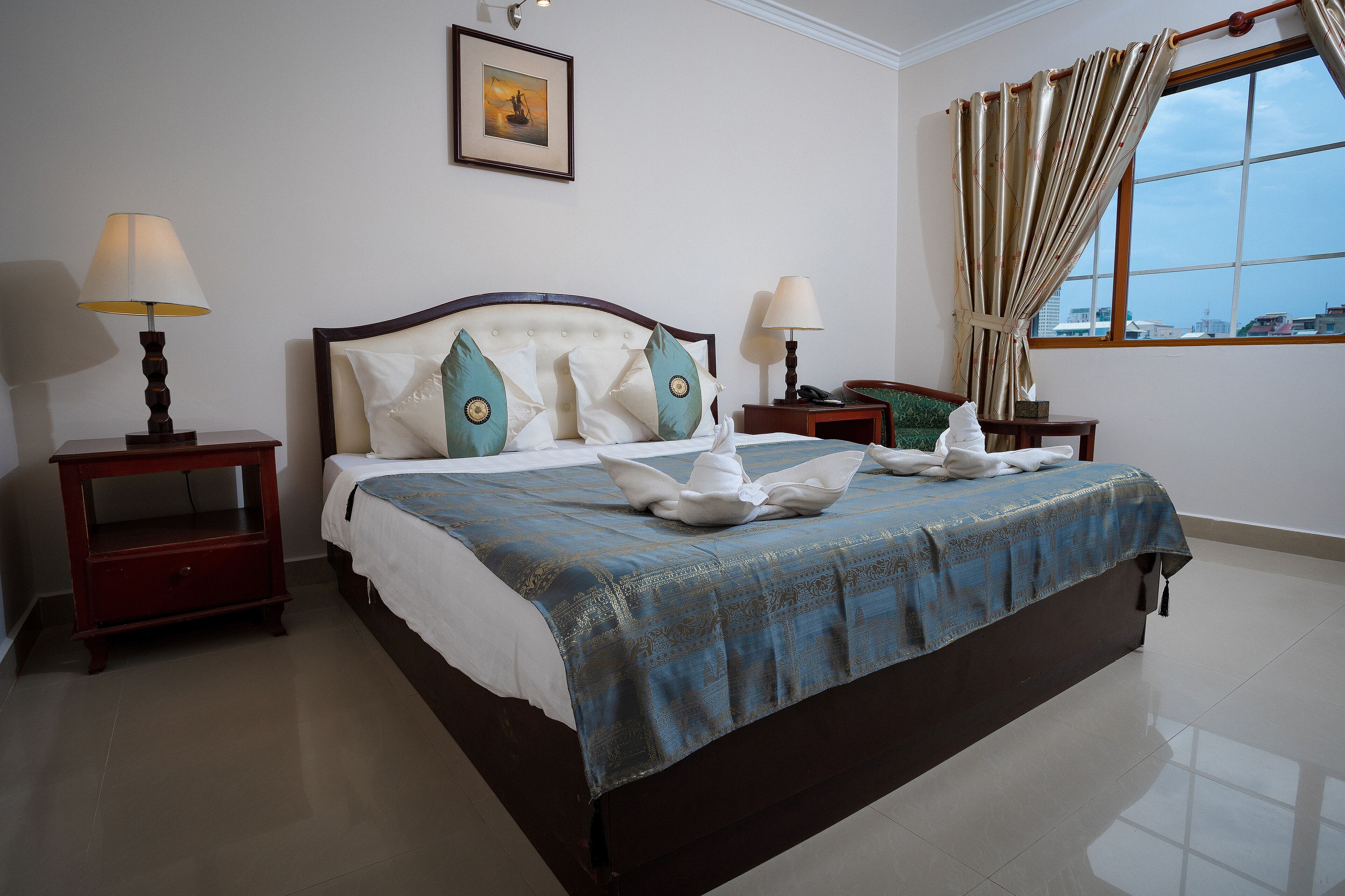 A neatly made bed with a blue bedspread, two bedside tables with lamps, a framed picture on the wall, and a window with curtains.