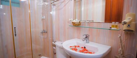 Un baño con cabina de ducha de vidrio, un lavamanos blanco adornado con pétalos de rosa, un estante de vidrio con artículos de aseo y una toalla en el inodoro.