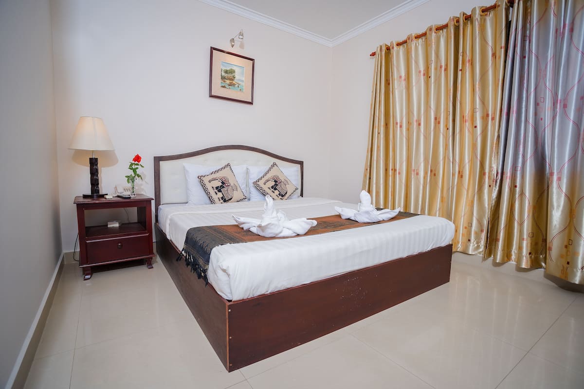 Una cama bien hecha con sábanas blancas y almohadones decorativos, una mesita de noche con una lámpara y un jarrón con una rosa roja, y un ventanal con cortinas.