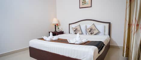 Una cama bien hecha con sábanas blancas y almohadas decorativas, una mesita de noche con una lámpara y un teléfono, y un cuadro en la pared.