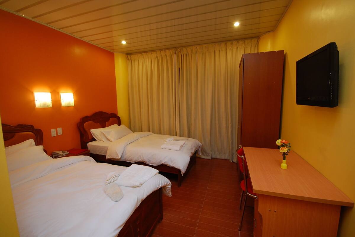 Ein Hotelzimmer mit zwei Betten, einem Holztisch, einem Stuhl, einem Fernseher und einem Fenster mit Vorhängen.