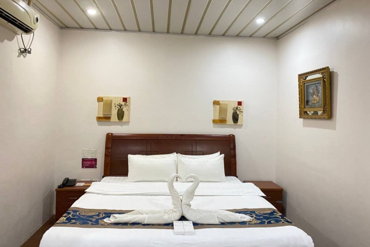 Una habitación de hotel con cabecera de madera, una cama bien hecha con ropa blanca y dos relojes en la pared.