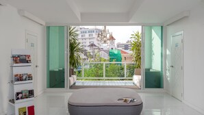 A modern room with a large window, a white sofa, a reading area with books, and a wicker ottoman. - Sino Imperial Design Hotel (Phuket)