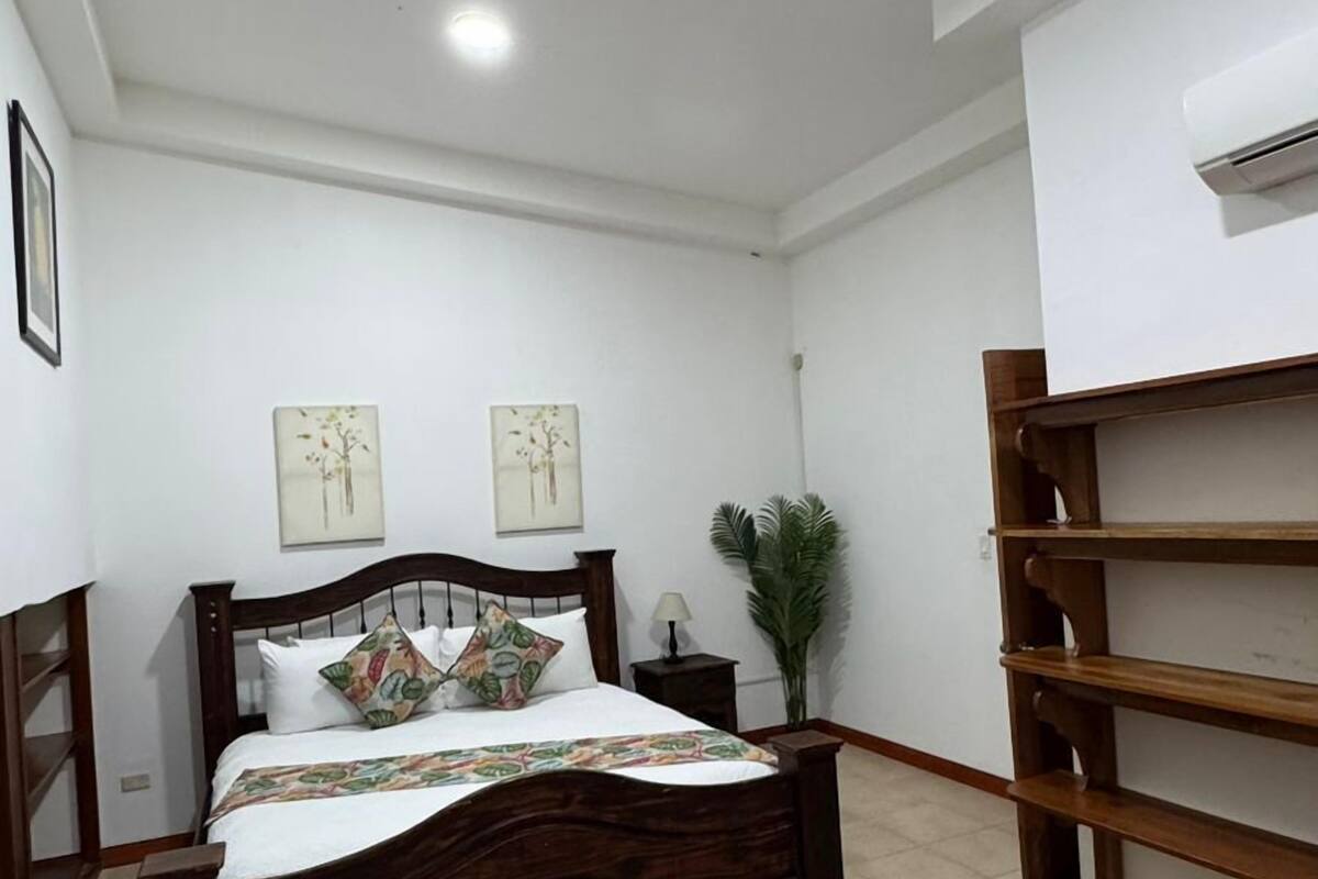 Un dormitorio con una cama de madera, una mesita de noche, aire acondicionado instalado en la pared y una lámpara en el techo.