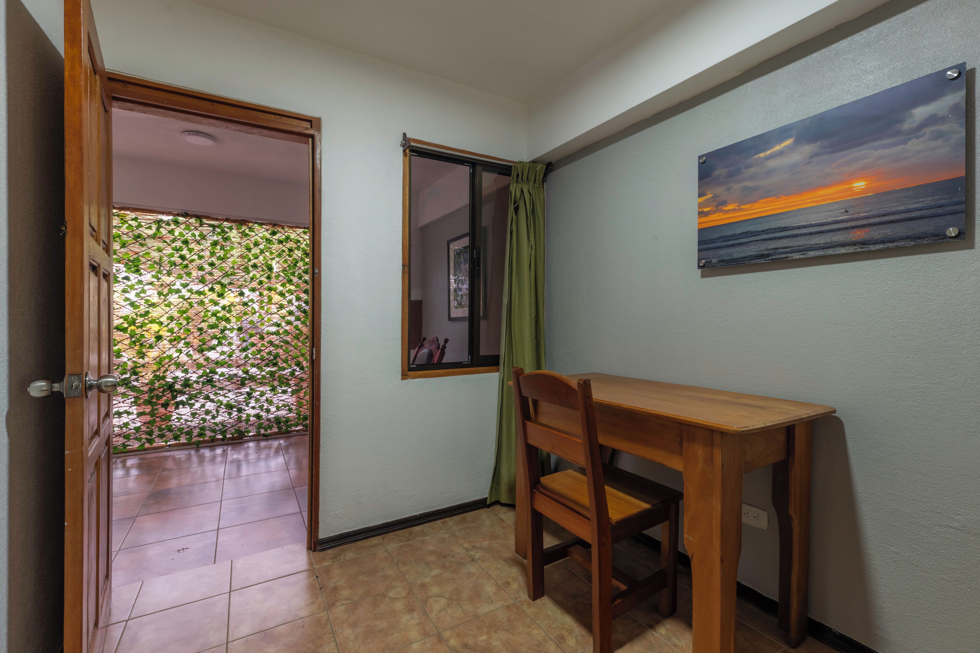 A room with a wooden table and chair, a window with a view of the sea, and a door leading to a balcony with a green plant wall.