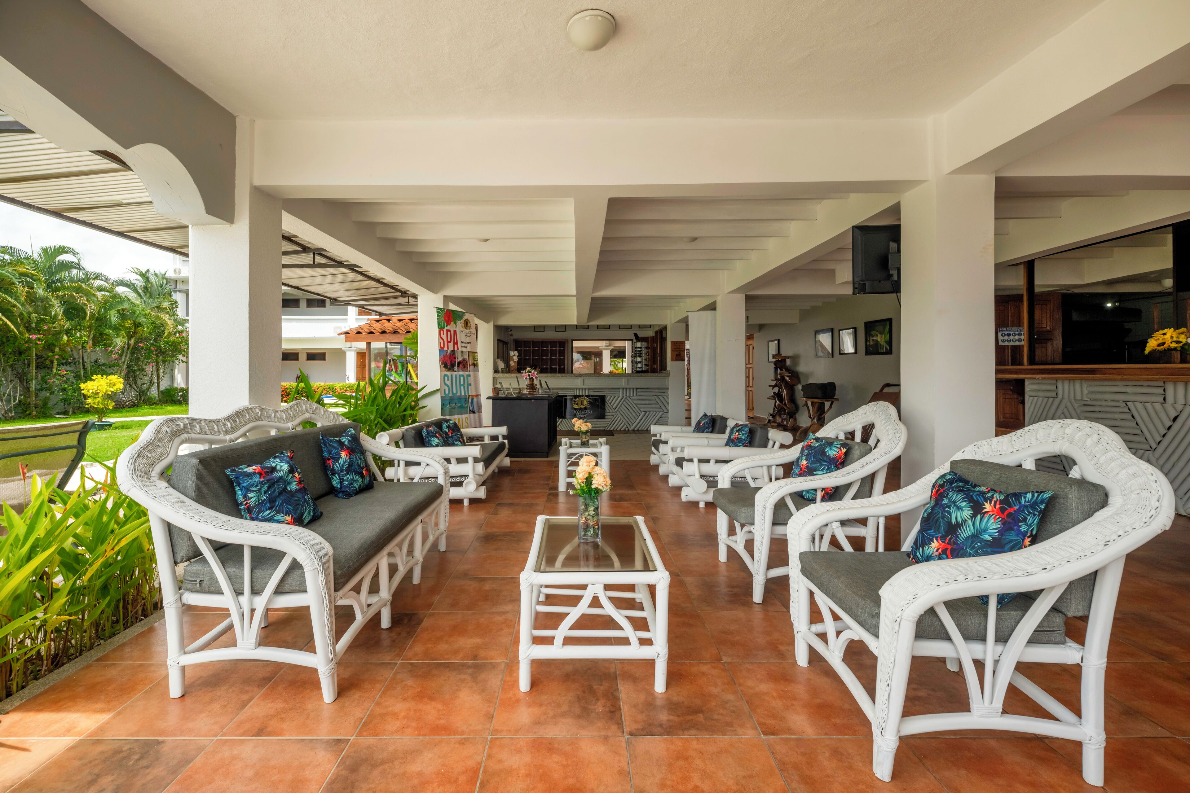 A covered outdoor seating area with wicker furniture and a central coffee table, surrounded by greenery and a view of the kitchen area.