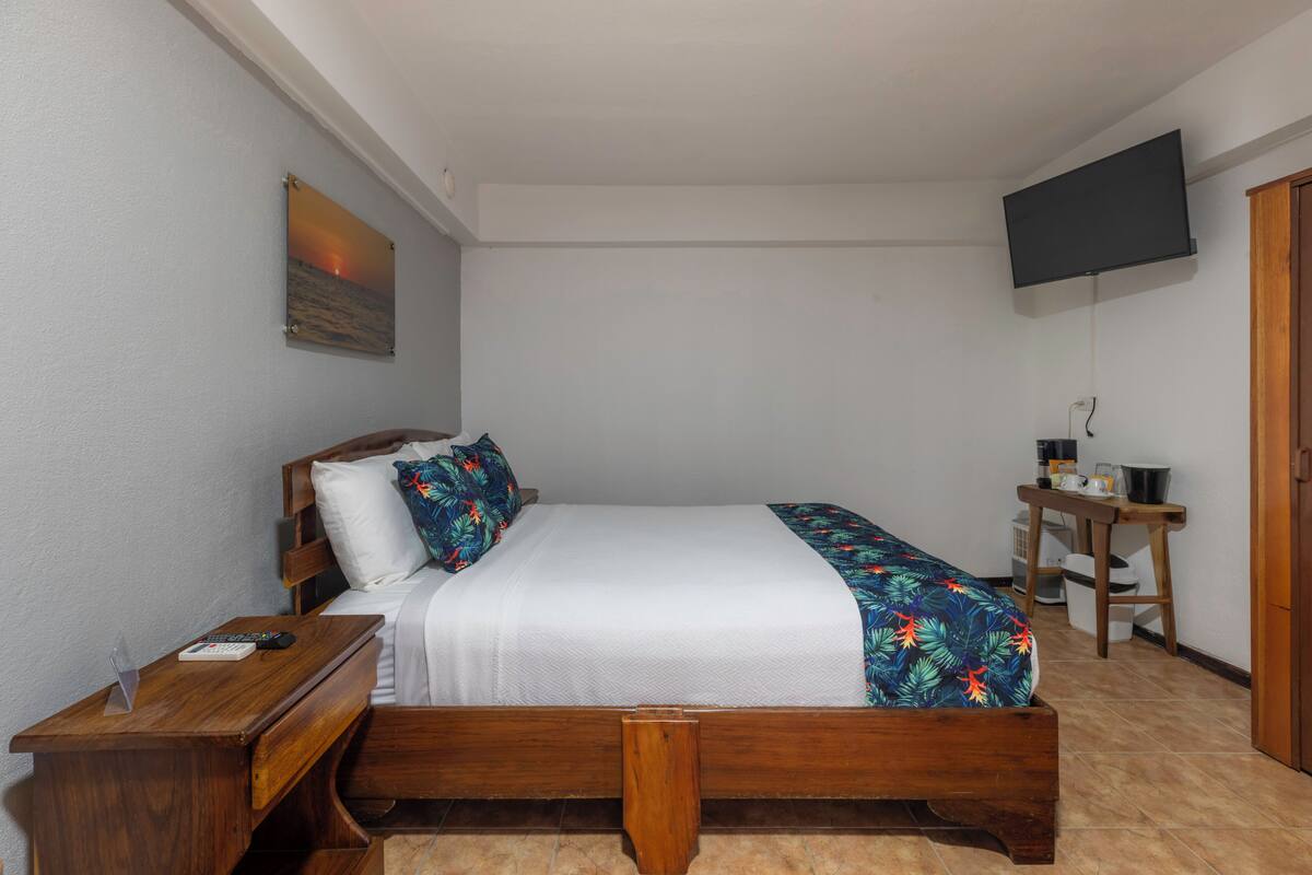 Un dormitorio con una cama, una mesita de noche de madera, un pequeño escritorio y una silla, y un televisor montado en la pared.