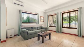 A living room with a green sofa, a wooden coffee table, and two windows with curtains.
