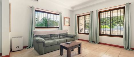 A living room with a green sofa, a wooden coffee table, and two windows with curtains.