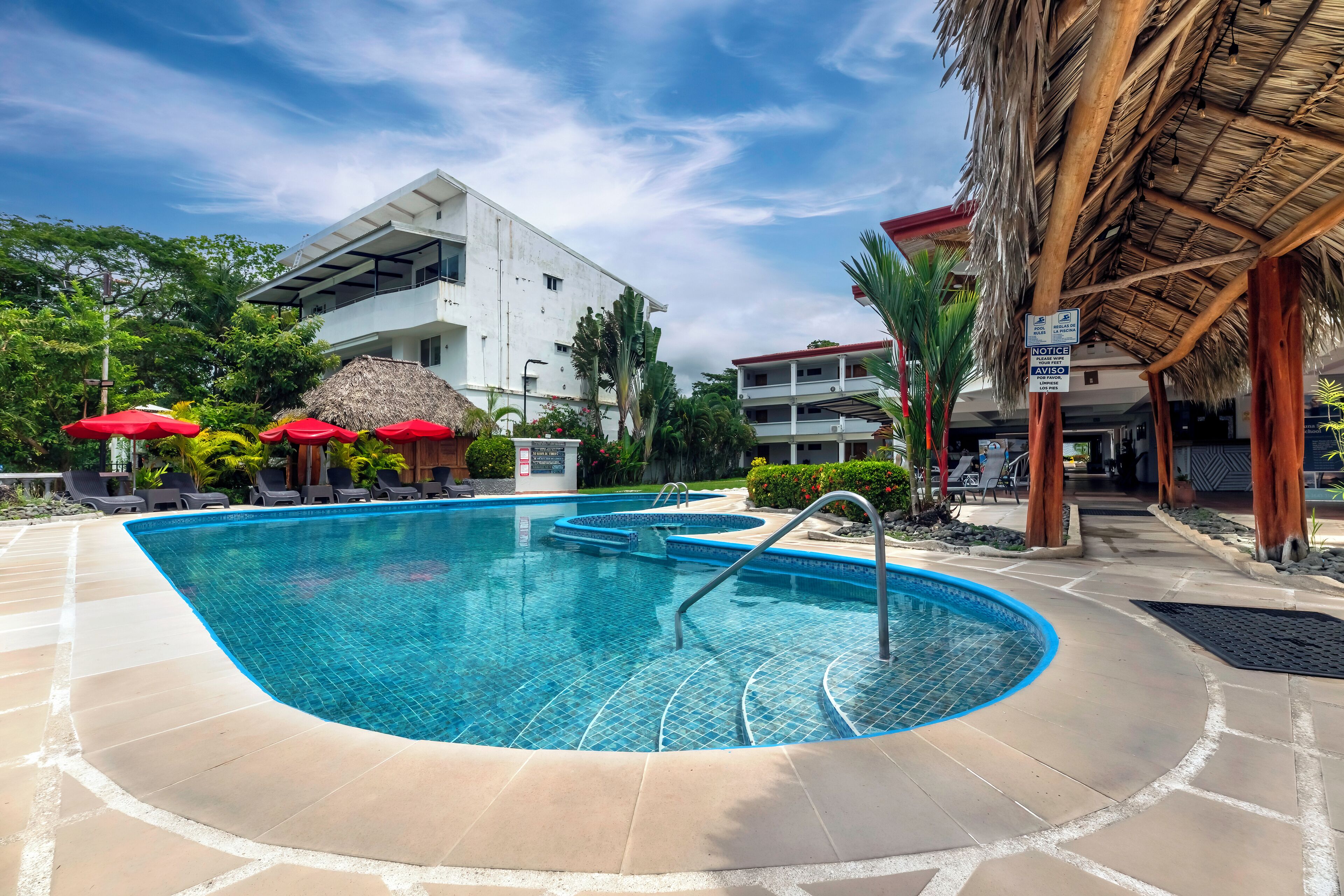 A swimming pool with a tiled deck, surrounded by lounge chairs and umbrellas, with a building in the background.