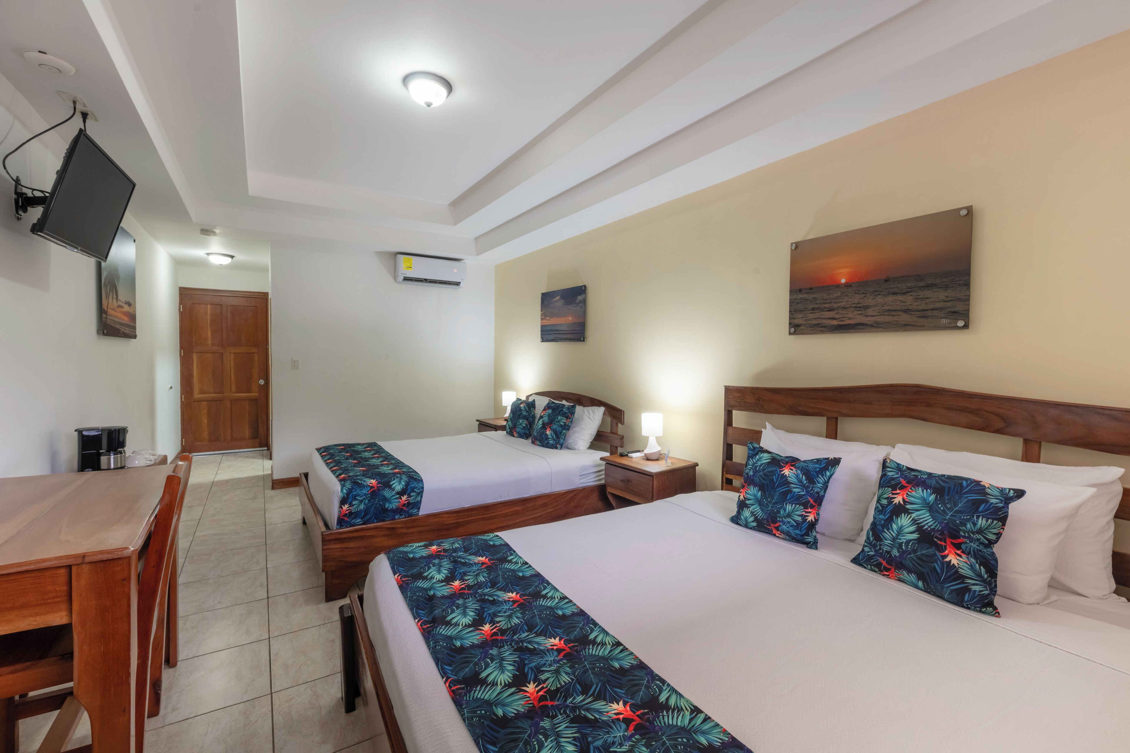 A hotel room with two beds, a wooden desk, a TV mounted on the wall, and a painting of a sunset.