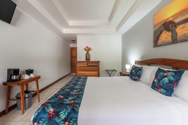 A hotel room with a bed, a wooden nightstand, a coffee maker, a trash bin, a wooden dresser, a vase with flowers, and a painting on the wall. - Jacó Laguna Resort and Beach Club (Jacó)