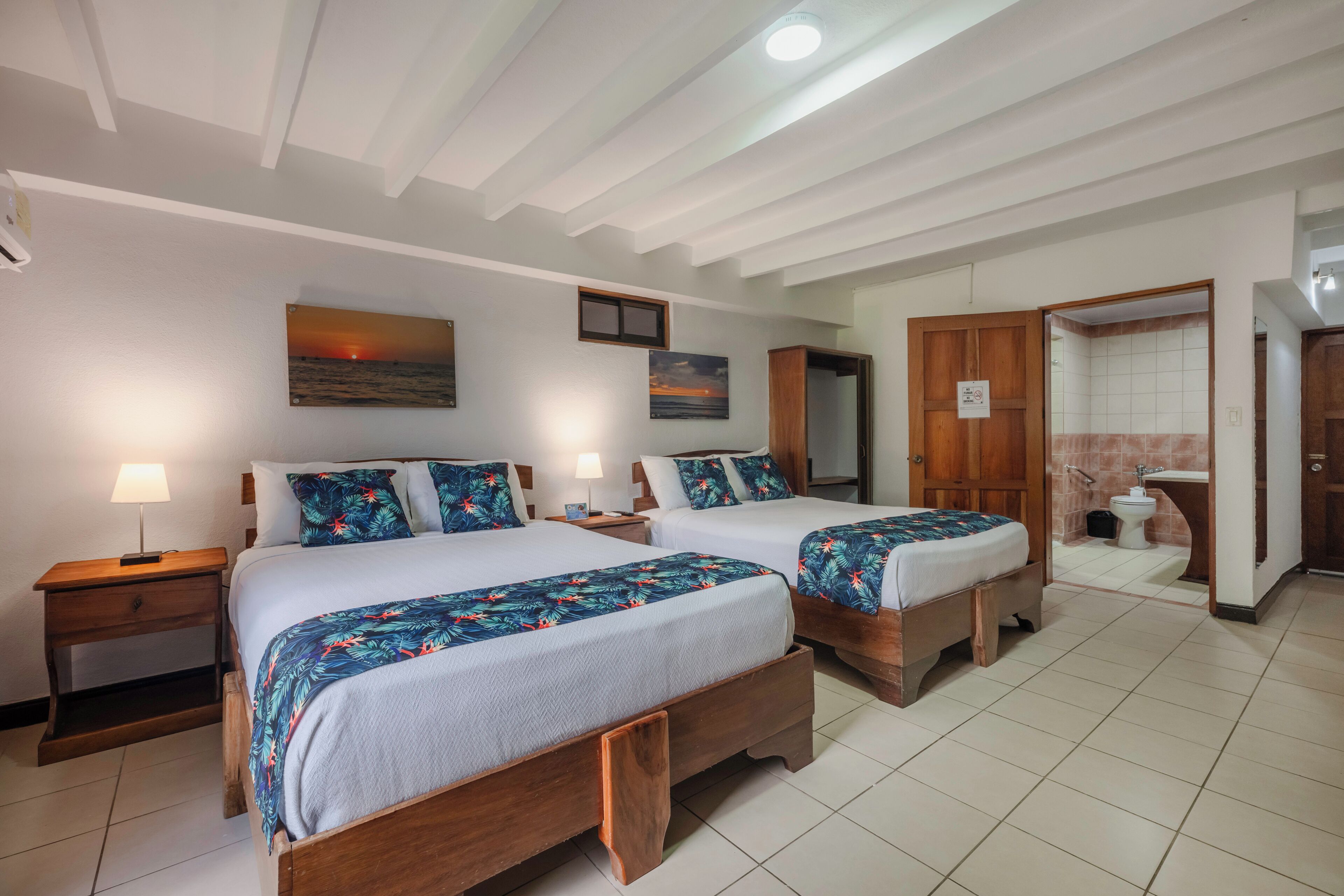 Quarto de hotel com duas camas de madeira, uma mesa de cabeceira, um quadro pendurado na parede e um banheiro visível através de uma porta aberta.