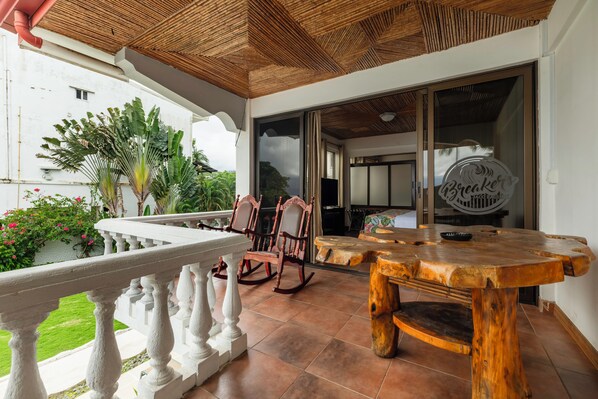 Un espacio al aire libre techado con una mesa de madera, mecedoras y una barandilla blanca.