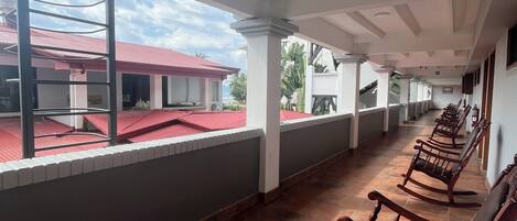 Área externa coberta com piso de azulejos, cadeiras de balanço de madeira e vista para um prédio com telhado vermelho.