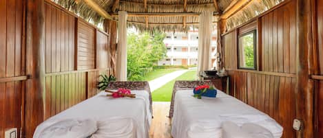 A room with two beds, a wooden ceiling, and a view of a building and greenery outside.